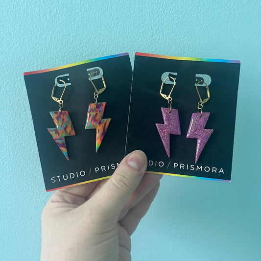 A hand holds two black display cards showing lightning bolt earrings against a light blue wall. One pair has a multicolor tie-dye pattern, and the other has a glittery purple finish.